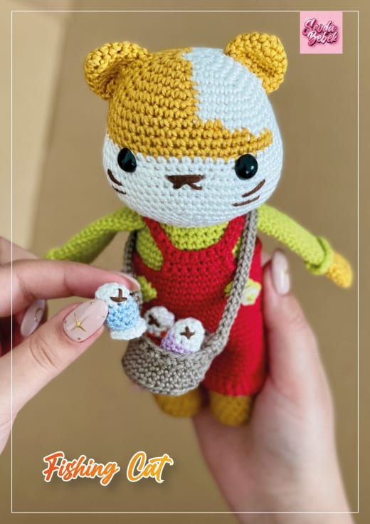 Crochet cat in a red outfit and green top holding a tiny basket with fish