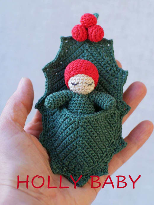 Tiny crochet baby with a red berry cap in a green holly leaf cradle held in a hand
