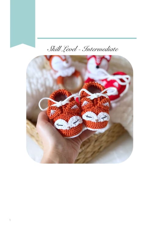 Pair of orange crochet baby booties with white fox faces held in a hand