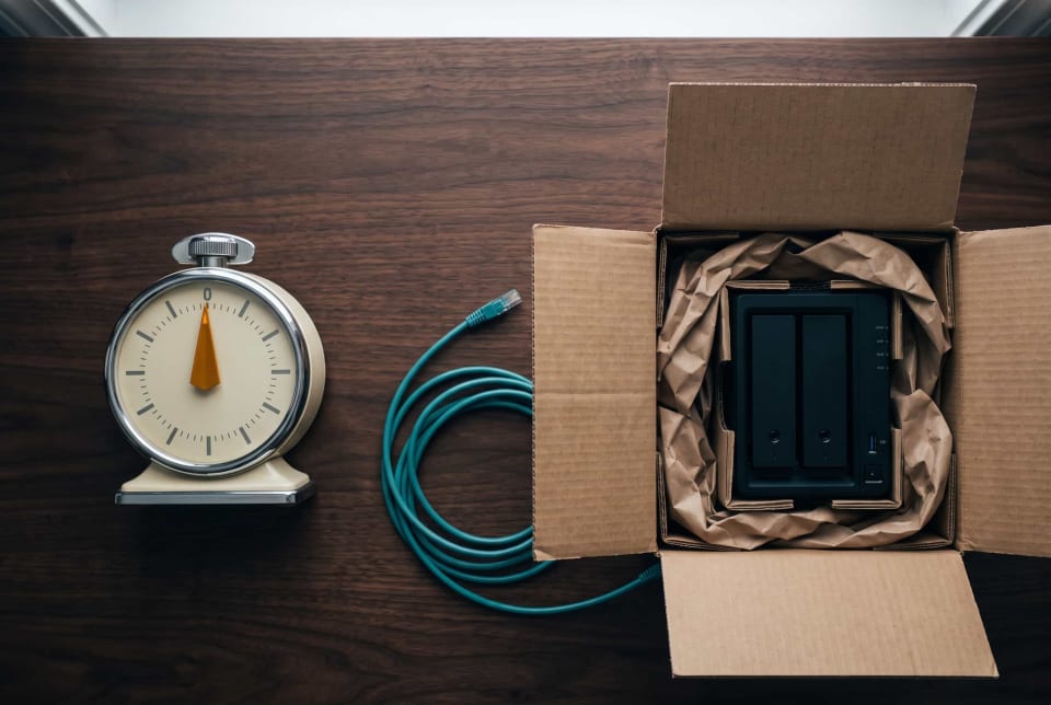 Wind-up kitchen timer beside an open shipping box holding a two-bay NAS on a walnut desk