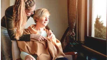 Journée mondiale des soins palliatifs 2025 : une jeune femme installe un châle autour d'une dame assise regardant au travers de la fenêtre.