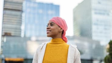 Chiffres clés du cancer du sein 2025 : tout savoir. Une jeune femme avec un foulard rose sur la tête dans une ville. Elle lance un regard plein d'espoir au loin.