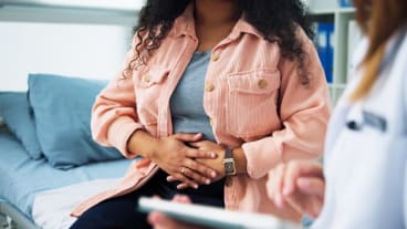 Endométriose : pourquoi cette maladie a longtemps été ignorée ? | ELSAN Actualités. Image d'une jeune femme dont on  ne voit pas le visage, qui se tient assise les mains sur le ventre devant une soignante en blouse blanche.