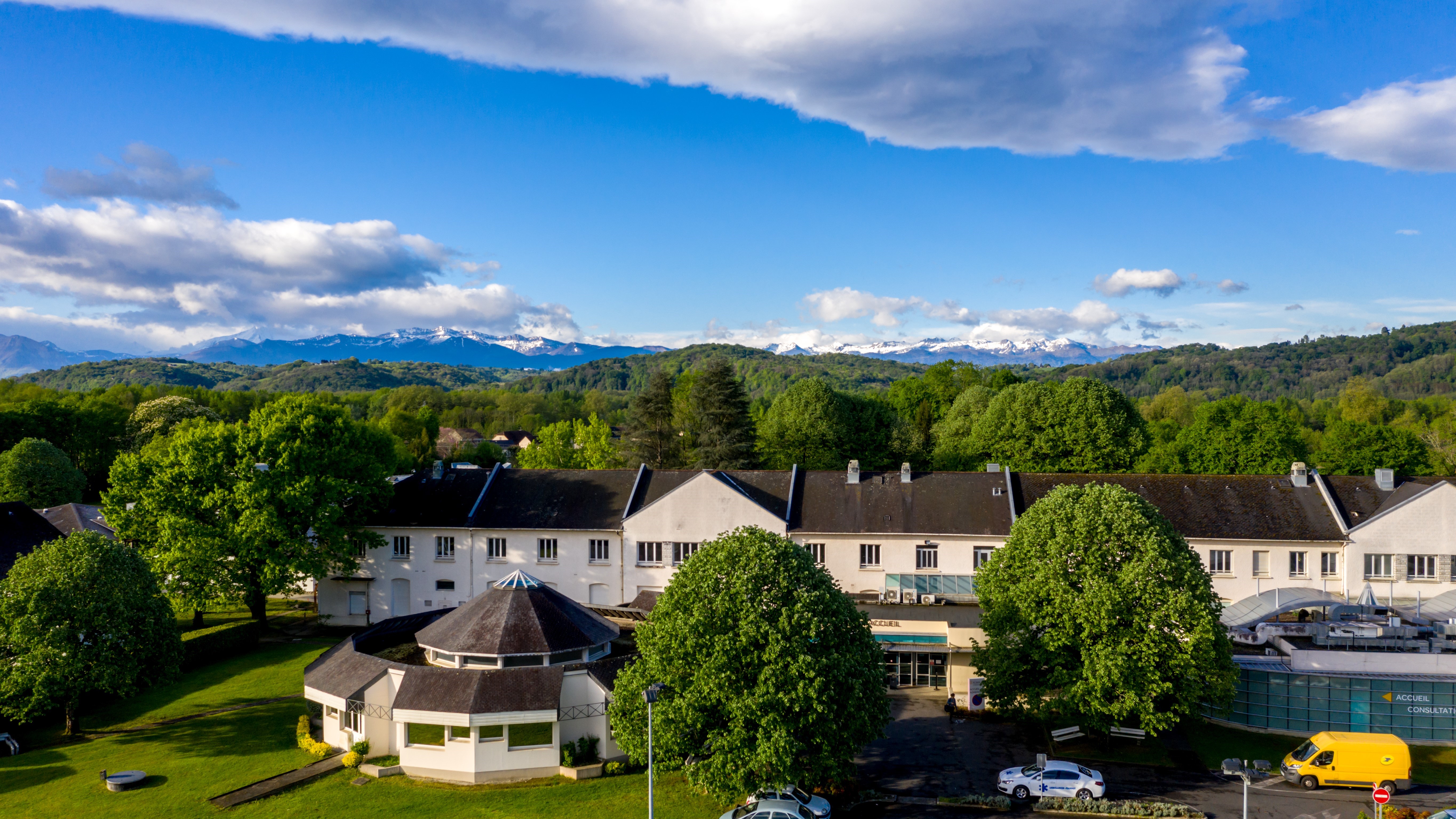 Aerial view of a healthcare facility surrounded by green trees, with mountains in the background under a partly cloudy blue sky.