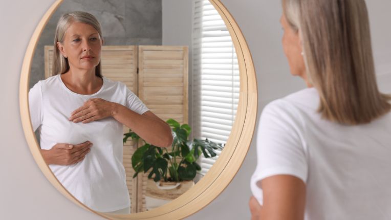 Cancer du sein : symptôme. Une femme s'autopalpe le sein au travers de son t-shirt devant le miroir.