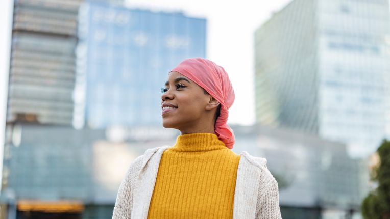 Chiffres clés du cancer du sein 2025 : tout savoir. Une jeune femme avec un foulard rose sur la tête dans une ville. Elle lance un regard plein d'espoir au loin.