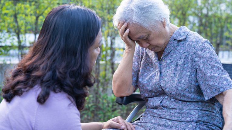 Infarctus du myocarde : définition, causes, traitement. Une personne âgée penche sa tête sur sa main. Une personne à son chevet s'inquiète de son état. 