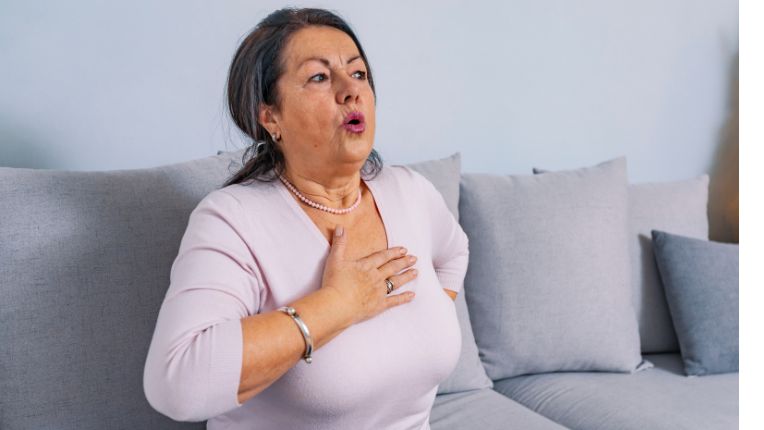Douleur thoracique chez la femme : symptômes crise cardiaque de la femme. Une femme d'environ 60 ans, se tient sur un sofa, avec des symptômes d'une possible crise cardiaque : visiblement angoissée, essoufflée, tentant de ventiler, avec la main droite sur le haut de la poitrine.
