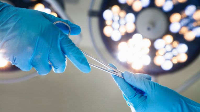 opération ganglions par robot Brest. Image de deux gants de soignants s'échangeant un outil chirurgical sous la lampe d'un champ opératoire.