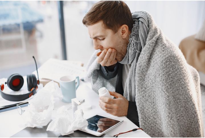 Symptômes de pneumonie : les reconnaître et réagir. Un jeune homme avec un plaid sur le dos tousse dans sa main et avale un cachet..
