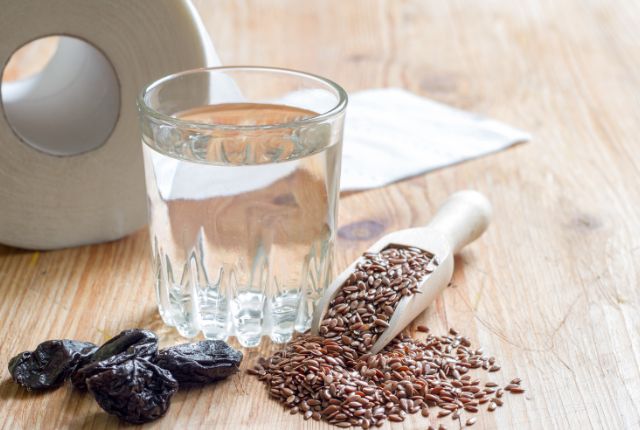 Constipation : table sur laquelle sont étalés des pruneaux, des graines, un verre d'eau et un rouleau de papier blanc.