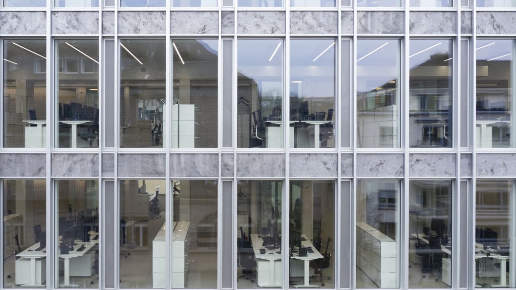 Bürofassade mit Glasfenstern, hinter denen helle Arbeitsplätze mit Schreibtischen sind.