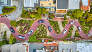 Luftaufnahme zeigt kurvenreiche, rot gepflasterte Straße mit mehreren Fahrzeugen, die zwischen Gebäuden verläuft
