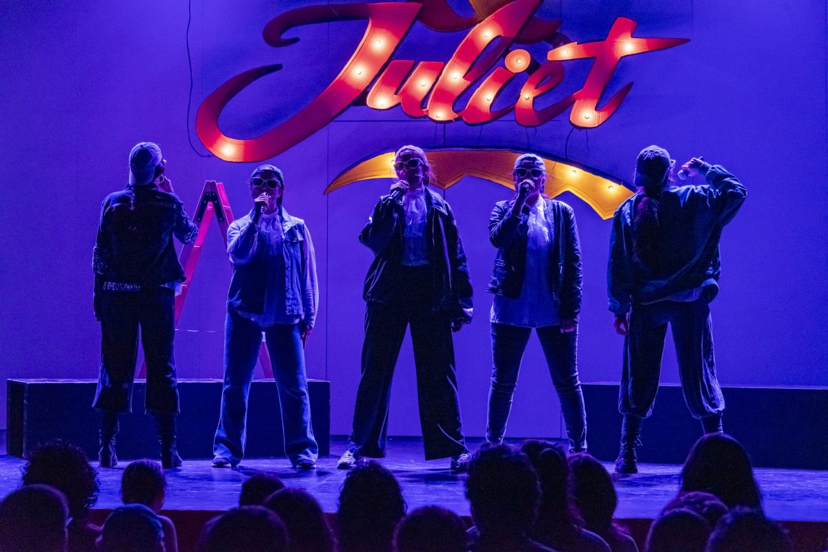 Seis intérpretes con cazadoras bajo un rótulo luminoso que dice «Juliet», con luz morada