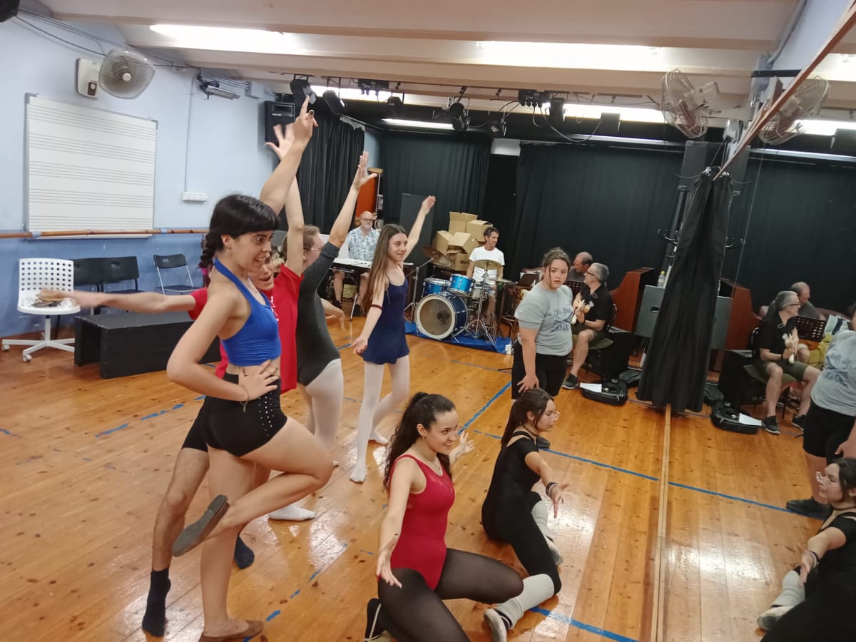 Ensayo con vestuario y batería al fondo, con espectadores sentados al lateral