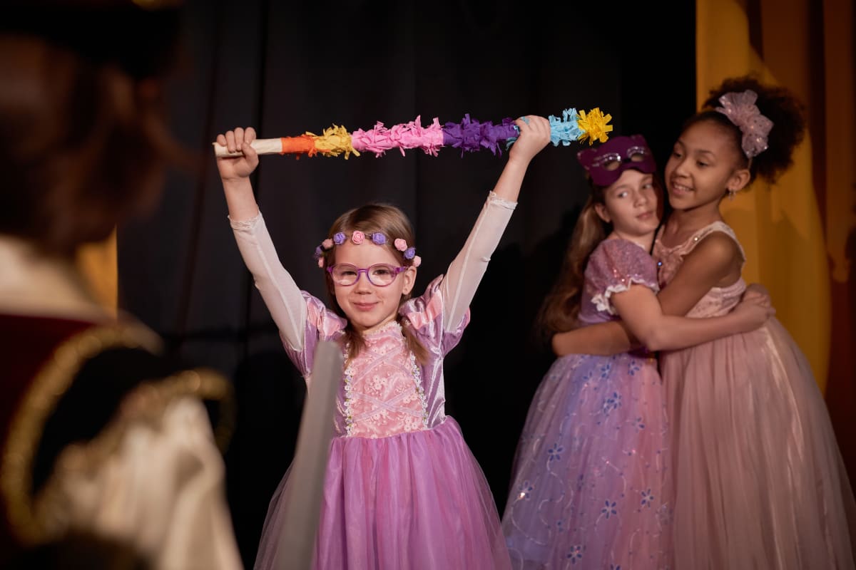Nena amb vestit de princesa aixeca una vareta de colors damunt l'escenari