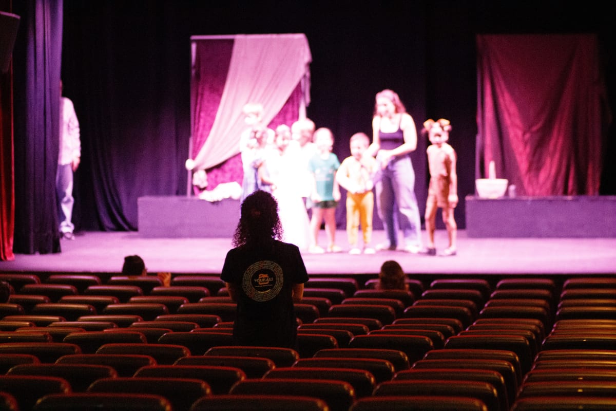 Ensayo visto desde el fondo de la sala vacía, con niños y una profesora en el escenario