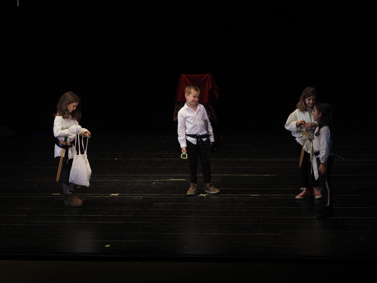 Quatre nens amb camisa blanca en un escenari ampli i fosc amb un tron al fons