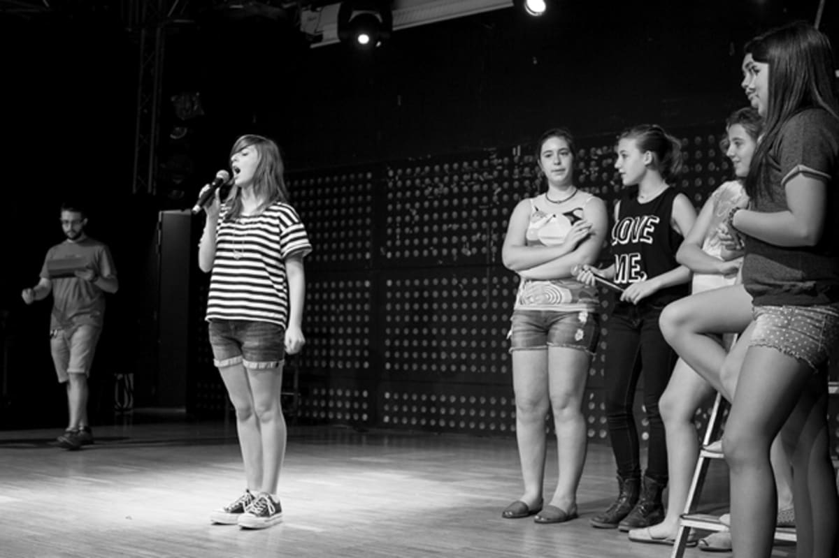 Foto en blanco y negro de una alumna cantando con micrófono mientras sus compañeras escuchan