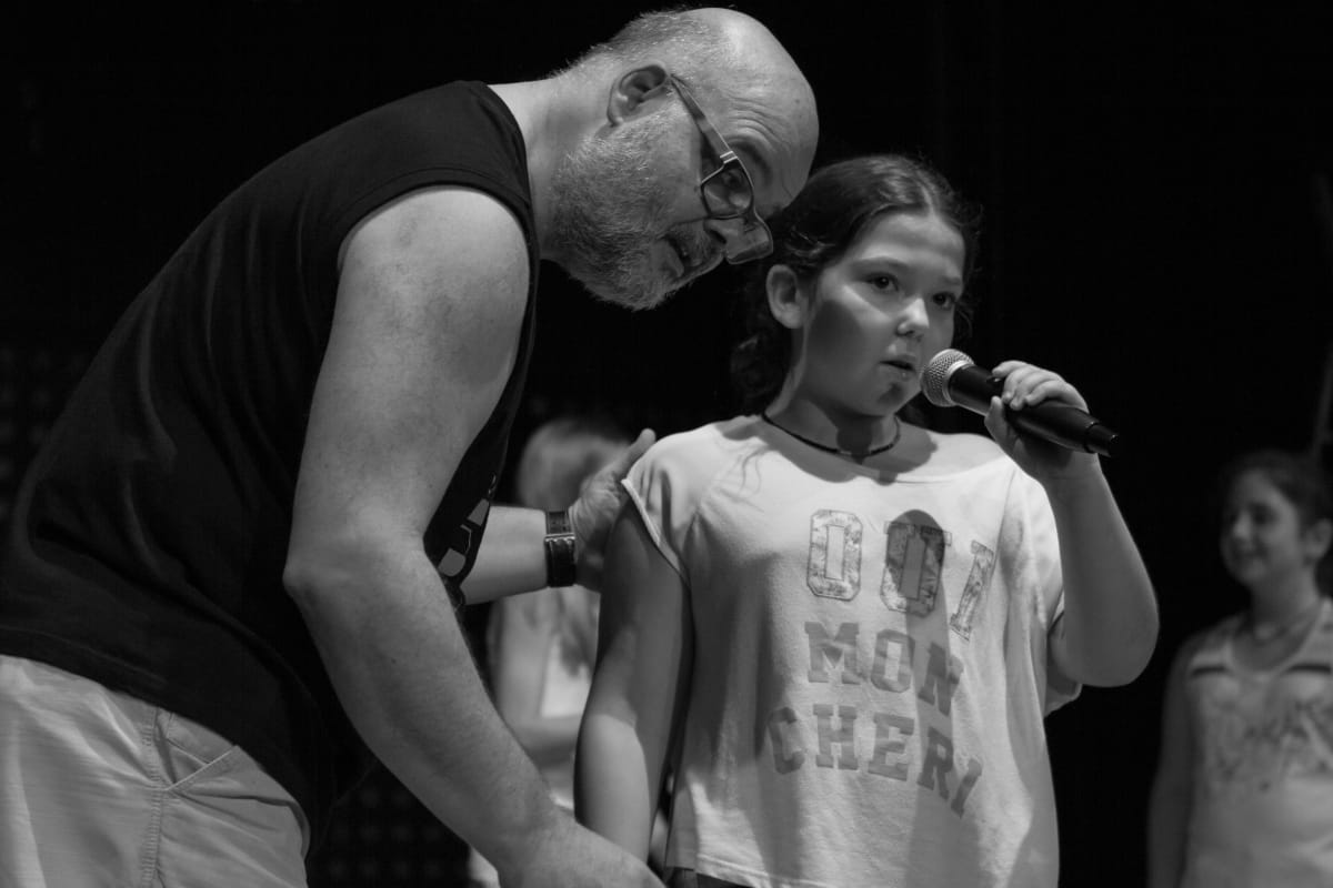 En blanco y negro, un profesor acompaña a una alumna que canta con micrófono