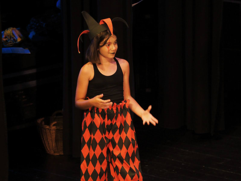 Nena amb barret de bufó i pantalons de rombes gesticulant sobre l'escenari