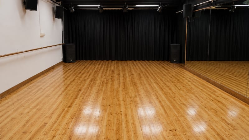 Aula vacía con parquet de madera, cortinas negras al fondo, pared de espejos, barra de danza y focos en el techo
