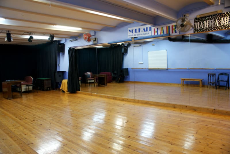 Sala de ensayo vacía con parquet, espejo de pared y carteles de musicales
