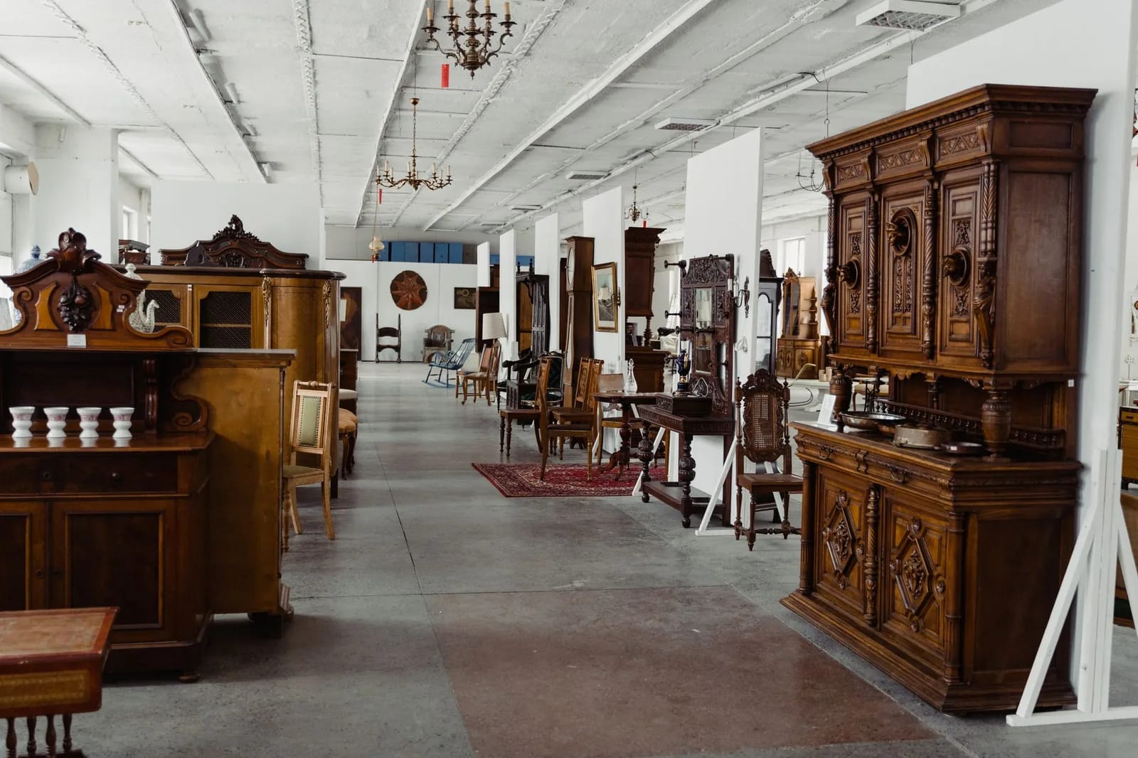 A long aisle of a multi-dealer antique showroom lined with carved wooden cabinets, chairs and chandeliers