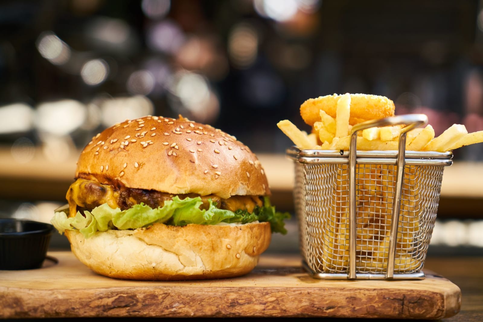 A cheeseburger on a wooden board beside a small wire basket of fries, against a dark blurred background
