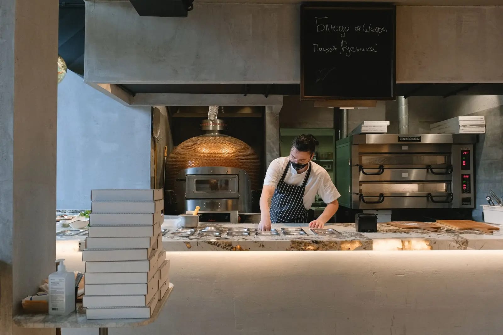 Masked chef preparing food in a modern pizzeria kitchen with ovens and pizza boxes