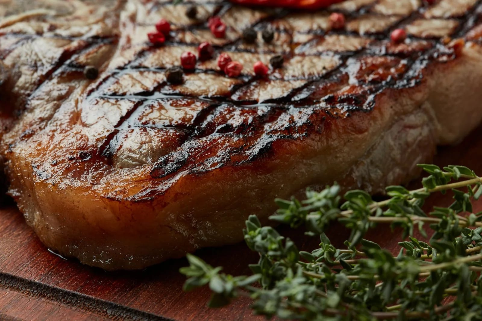A thick grilled steak with cross-hatched grill marks, peppercorns and fresh thyme on a wooden board
