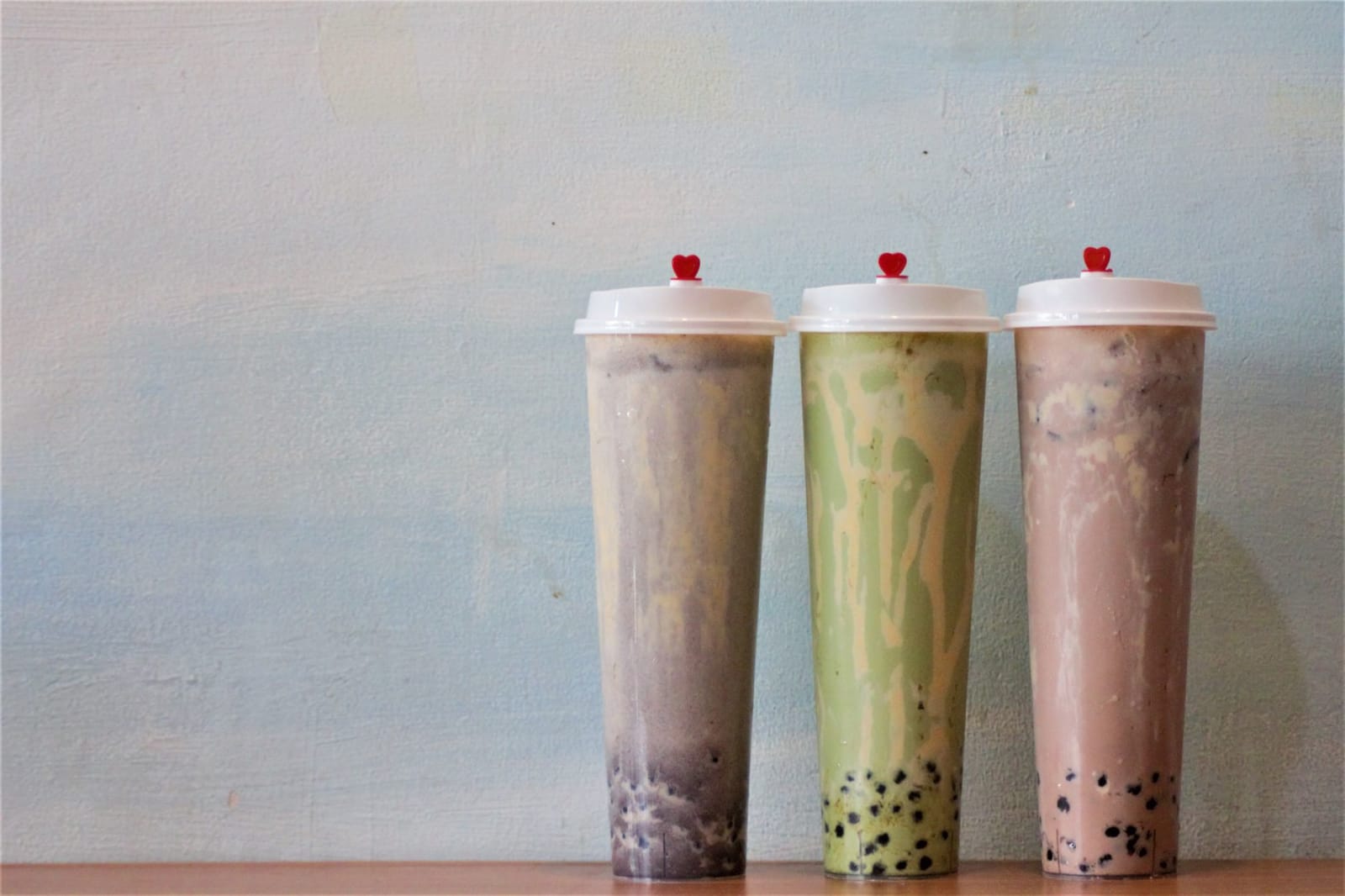 Three tall sealed cups of bubble tea in different colors lined up against a pale wall, dark tapioca pearls settled in each base