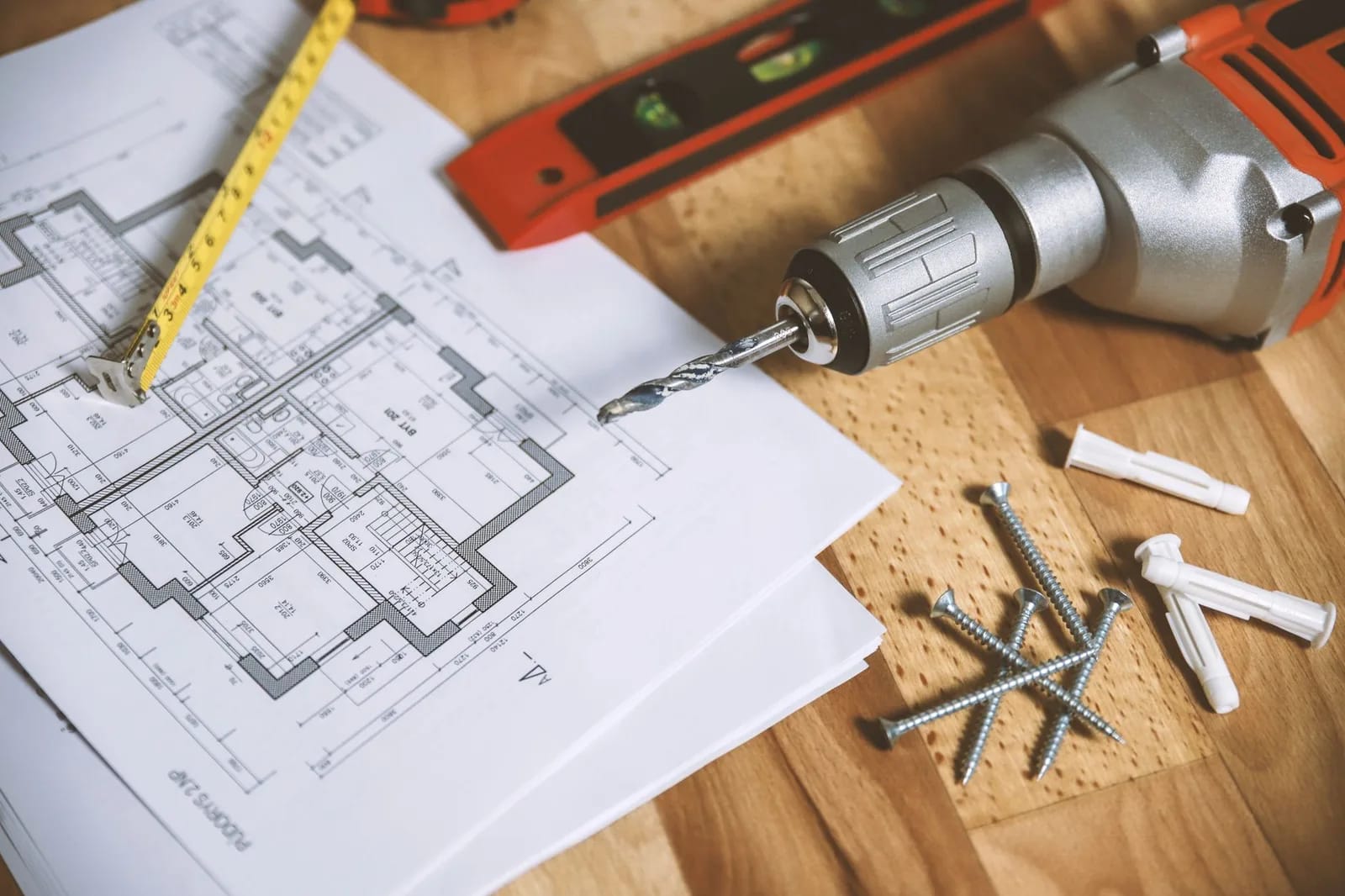 A residential floor plan on a wooden floor beside a tape measure, a level, a cordless drill, screws and wall anchors