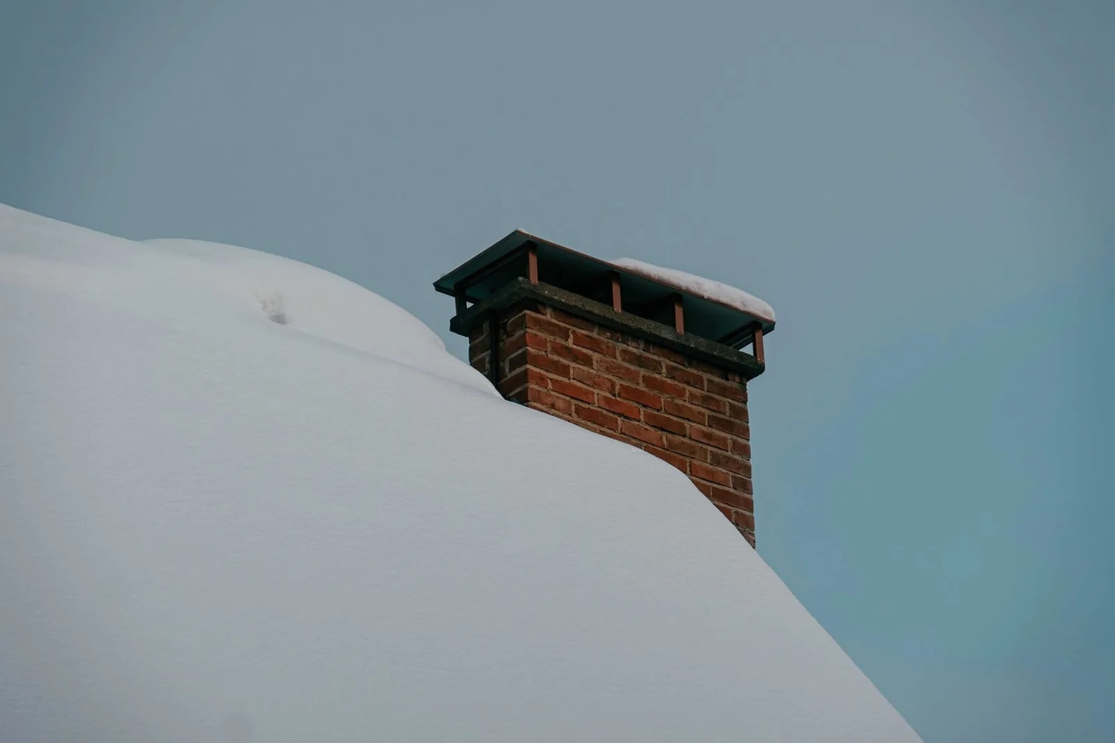 A red brick chimney topped with a metal rain cap, rising from a roof under deep snow