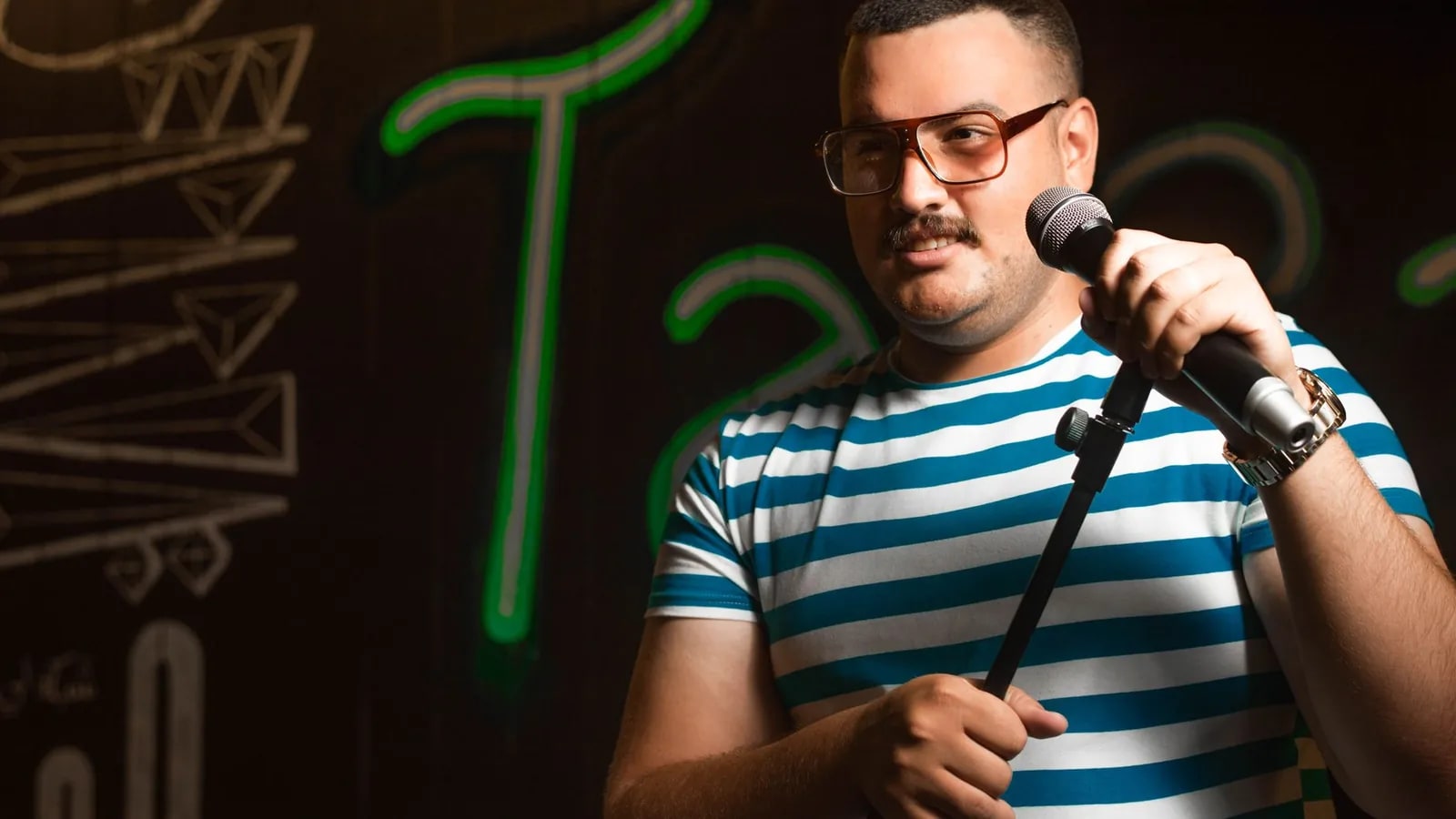 A stand-up comedian holding a microphone on stage in front of a neon-lit wall