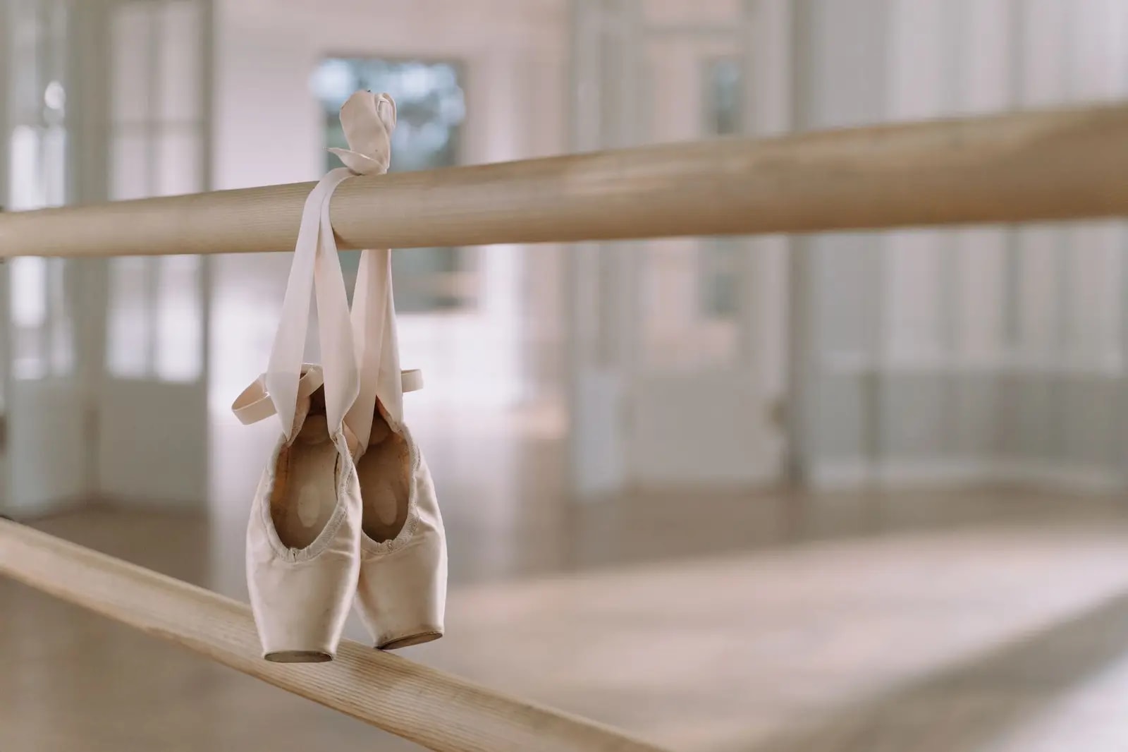 A pair of well-worn pointe shoes hanging by their ribbons from a wooden ballet barre in a bright studio