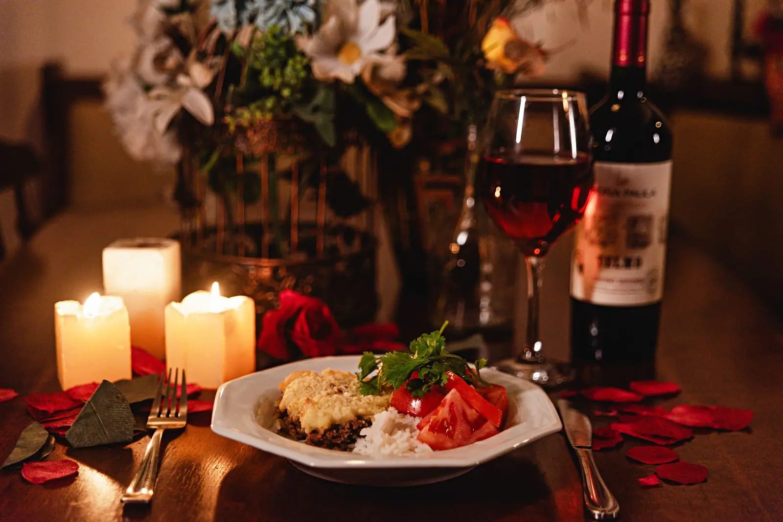 Cozy candlelit dinner featuring wine, a gourmet meal, and romantic decor with rose petals
