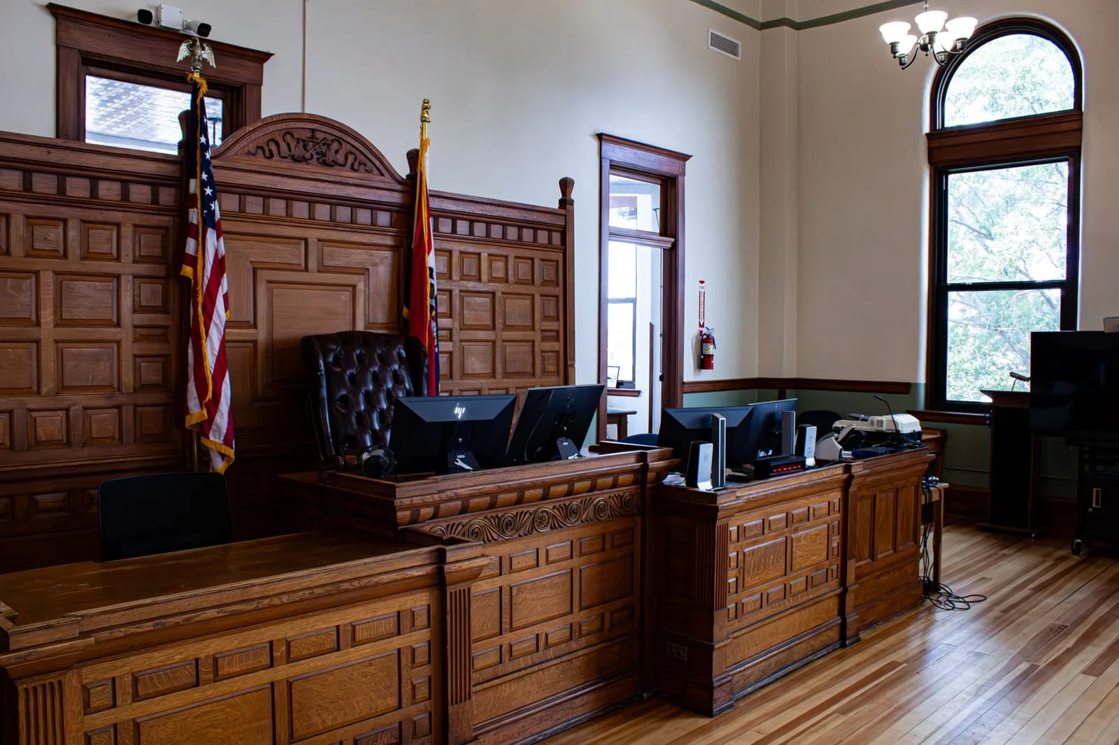 A wood-paneled courtroom with a leather judge's chair, computer monitors on the bench, and the United States flag beside a state flag