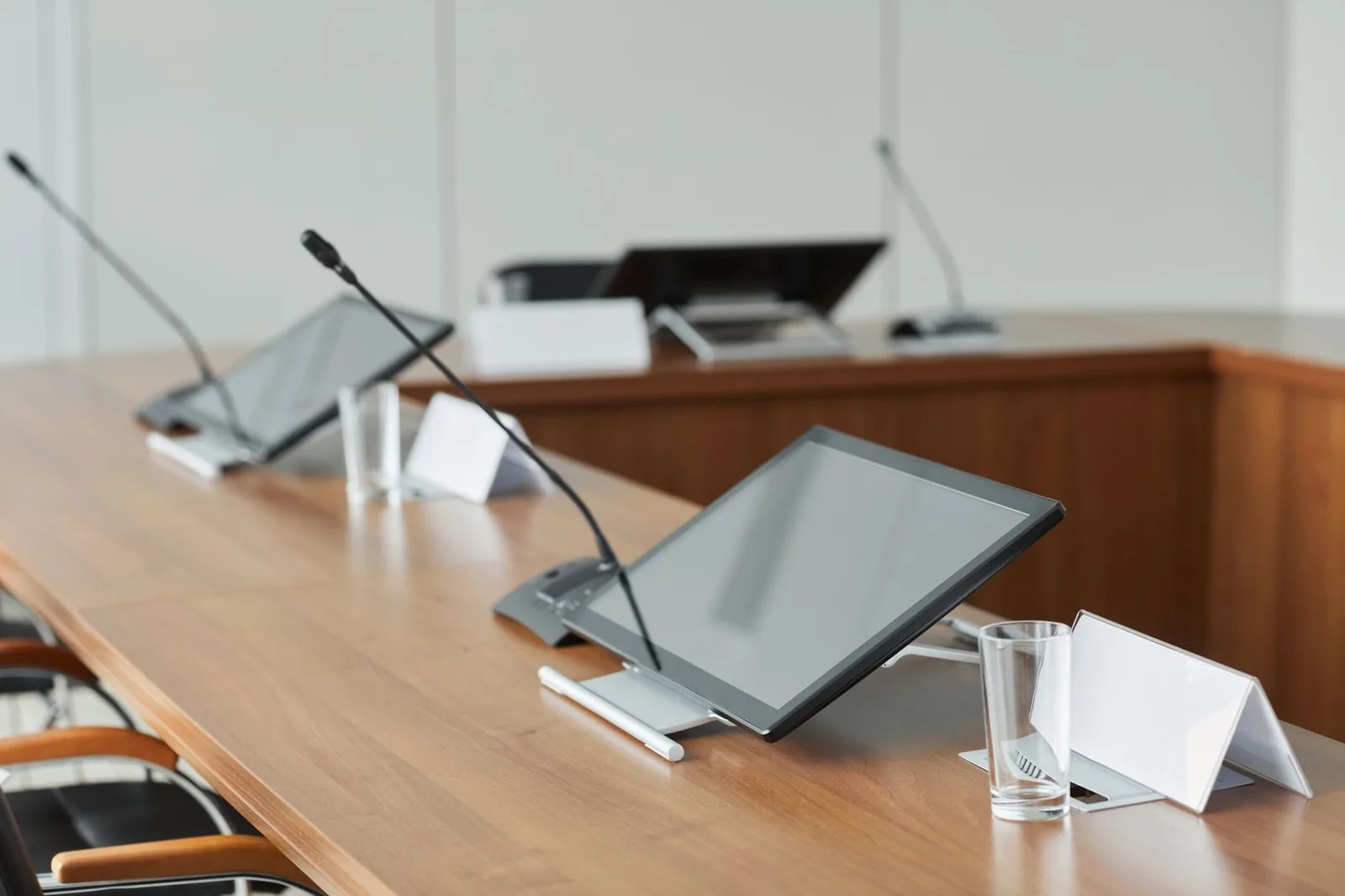 Microphones and tablet screens set along a polished wooden meeting table with blank name-card holders and a water glass