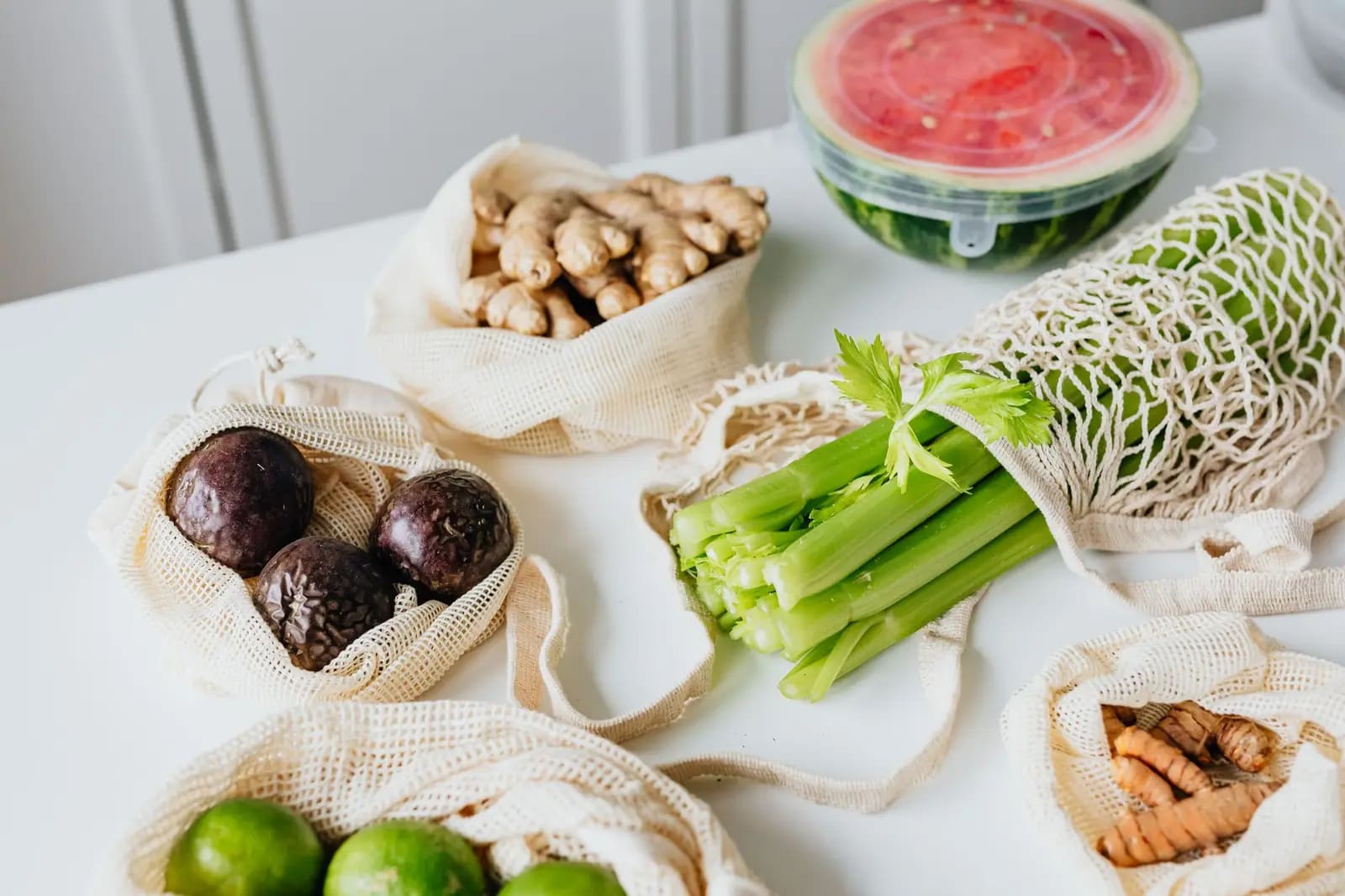 Fresh vegetables and fruit in reusable mesh bags arranged on a white table