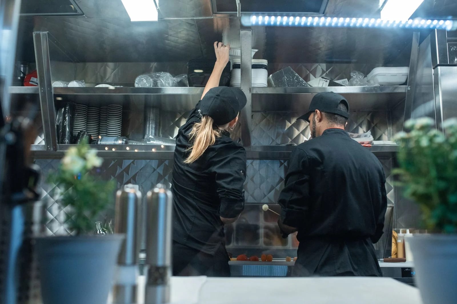 Two cooks in black jackets and caps working at a stainless steel prep counter in a commercial kitchen