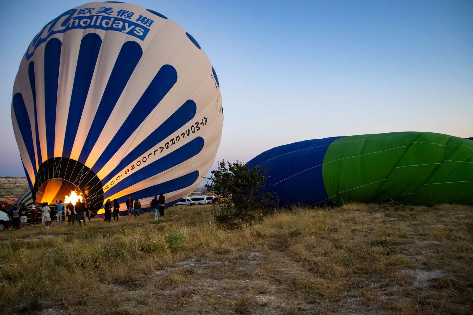 A hot air balloon standing upright at dawn with its burner lit, a second envelope still lying inflated on the grass beside it