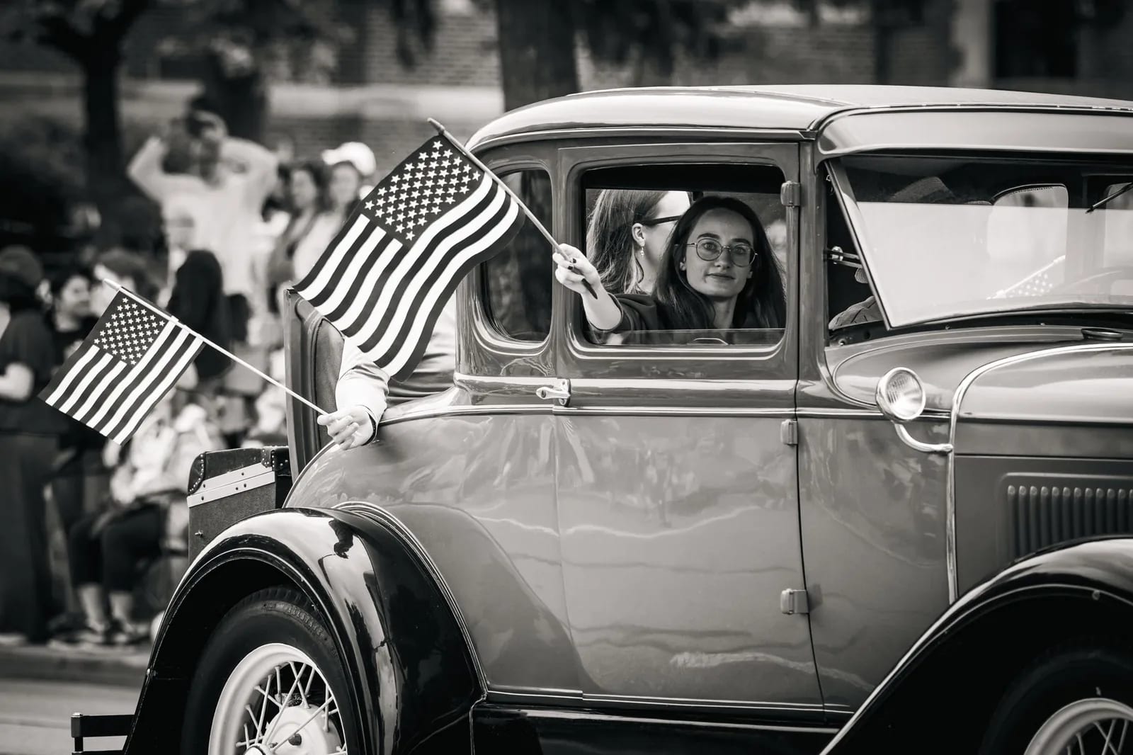 A classic car in a parade with American flags held up along the route