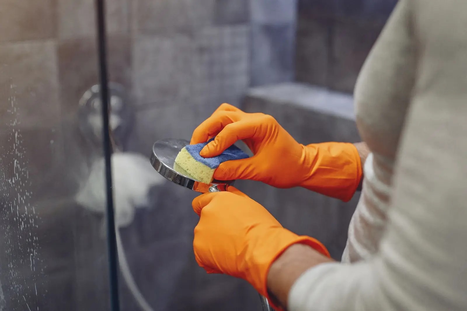 Gloved hands cleaning a shower head with a sponge inside a glass shower enclosure