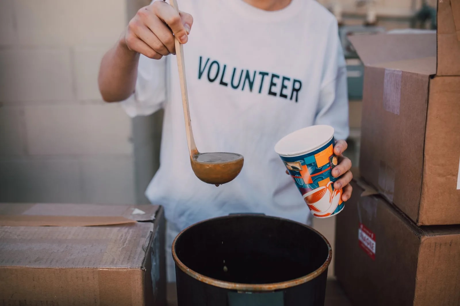 A volunteer in a lettered T-shirt lifting a ladle over a soup pot beside stacked cardboard boxes