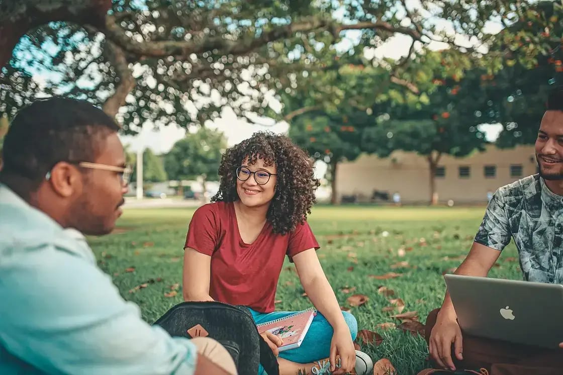 Three students sitting on a campus lawn with a laptop and books