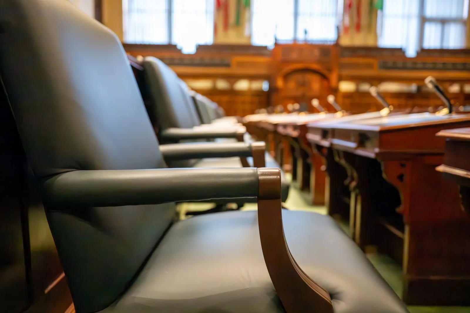 Green leather seats with wooden armrests in the foreground of a wood-paneled chamber, with microphones on desks beyond