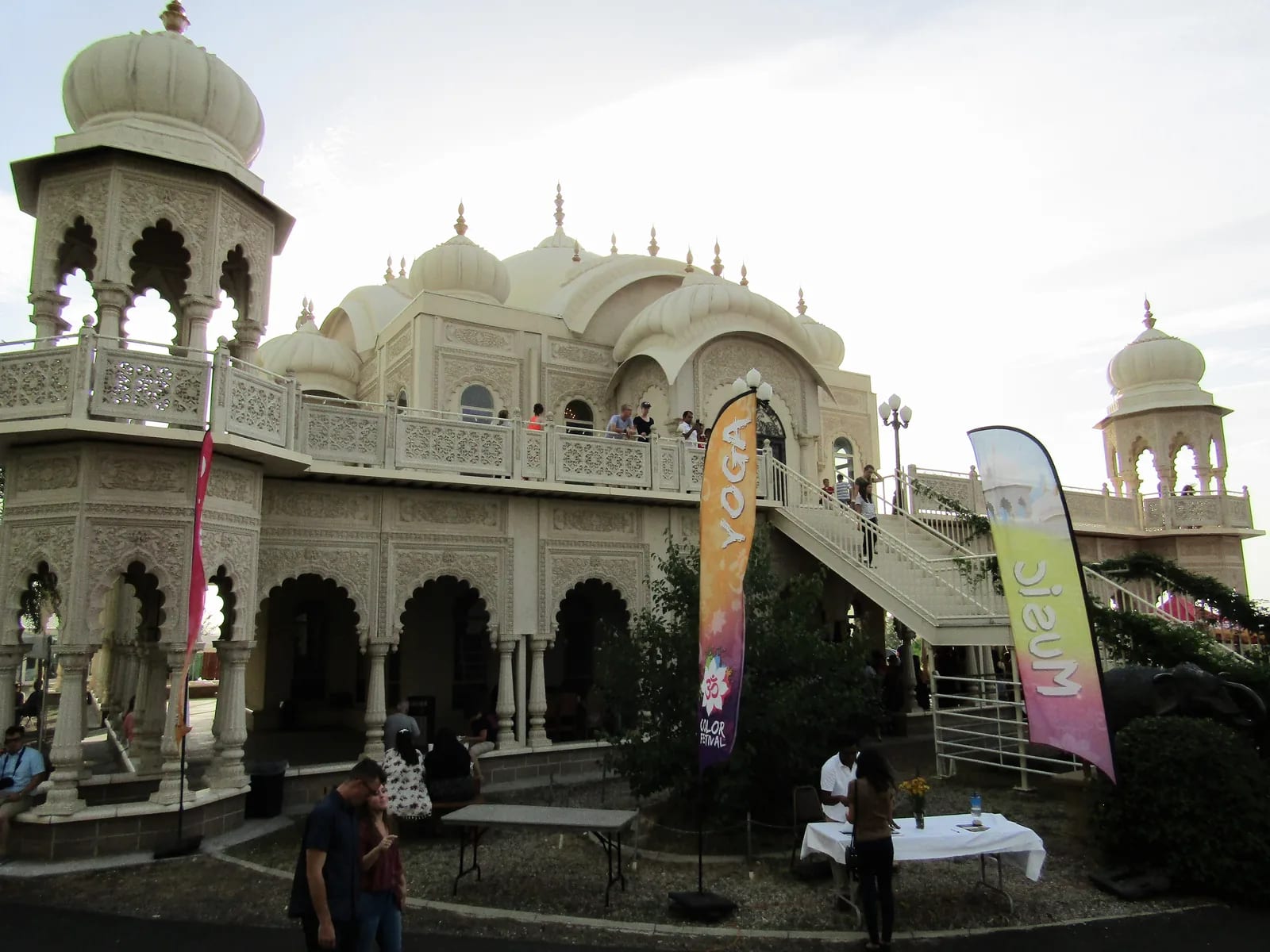 A white domed temple with carved arches and balconies, with event banners and visitors on the steps