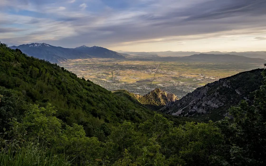 The south end of Utah Valley seen from the road below Kyhv Peak, with forested slopes falling away and the Wasatch ridgeline receding into haze