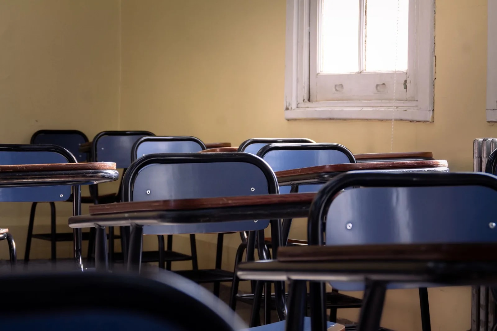 Rows of empty blue tablet-arm chairs in a classroom lit by a single window